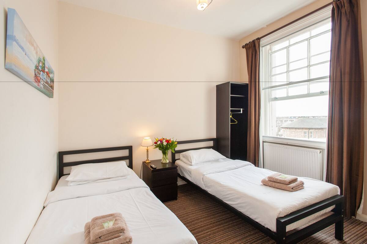 Ett hotellrum med två separata sängar, ett nattduksbord med en lampa och blommor, en garderob och ett fönster med gardiner.