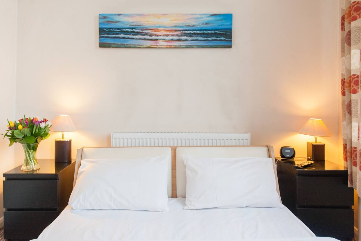 Ett hotellrum med en säng, två sängbord, ett litet bord, en lampa, en vas med blommor och en tavla som föreställer en solnedgång över havet.