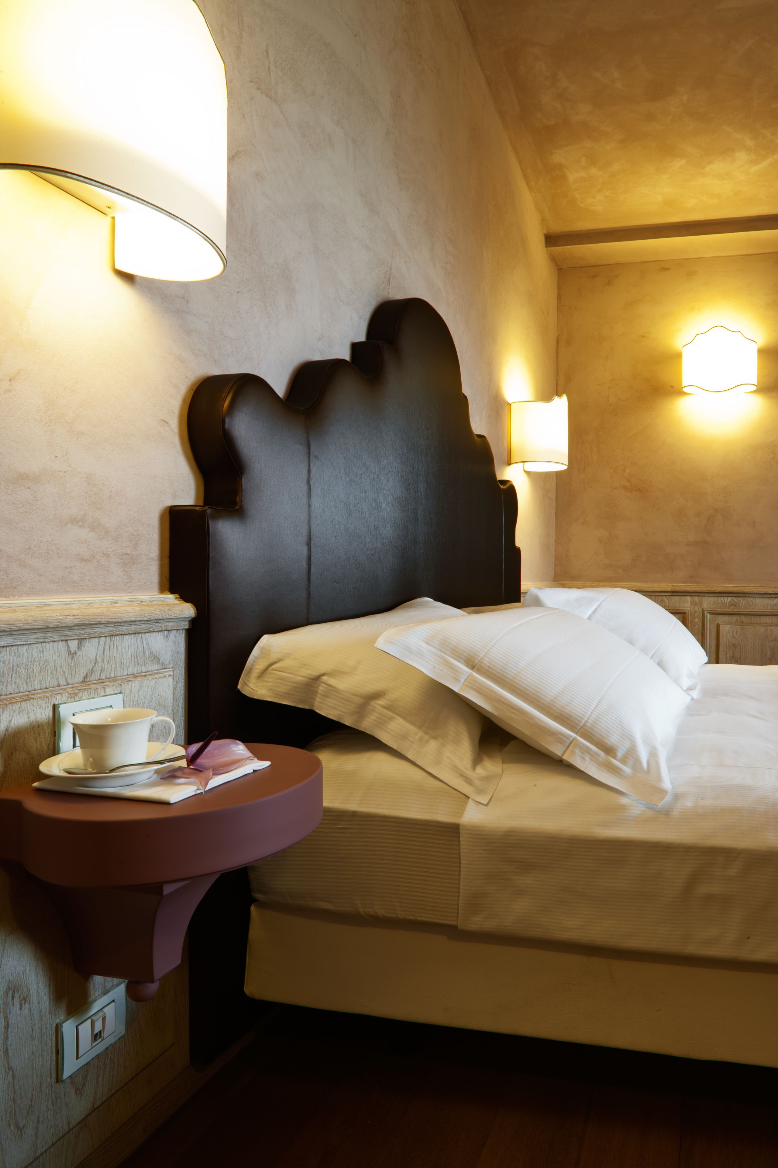 Une chambre d’hôtel avec un lit, des lampes de chevet, une petite table avec une tasse et un livre, et un luminaire fixé au mur.