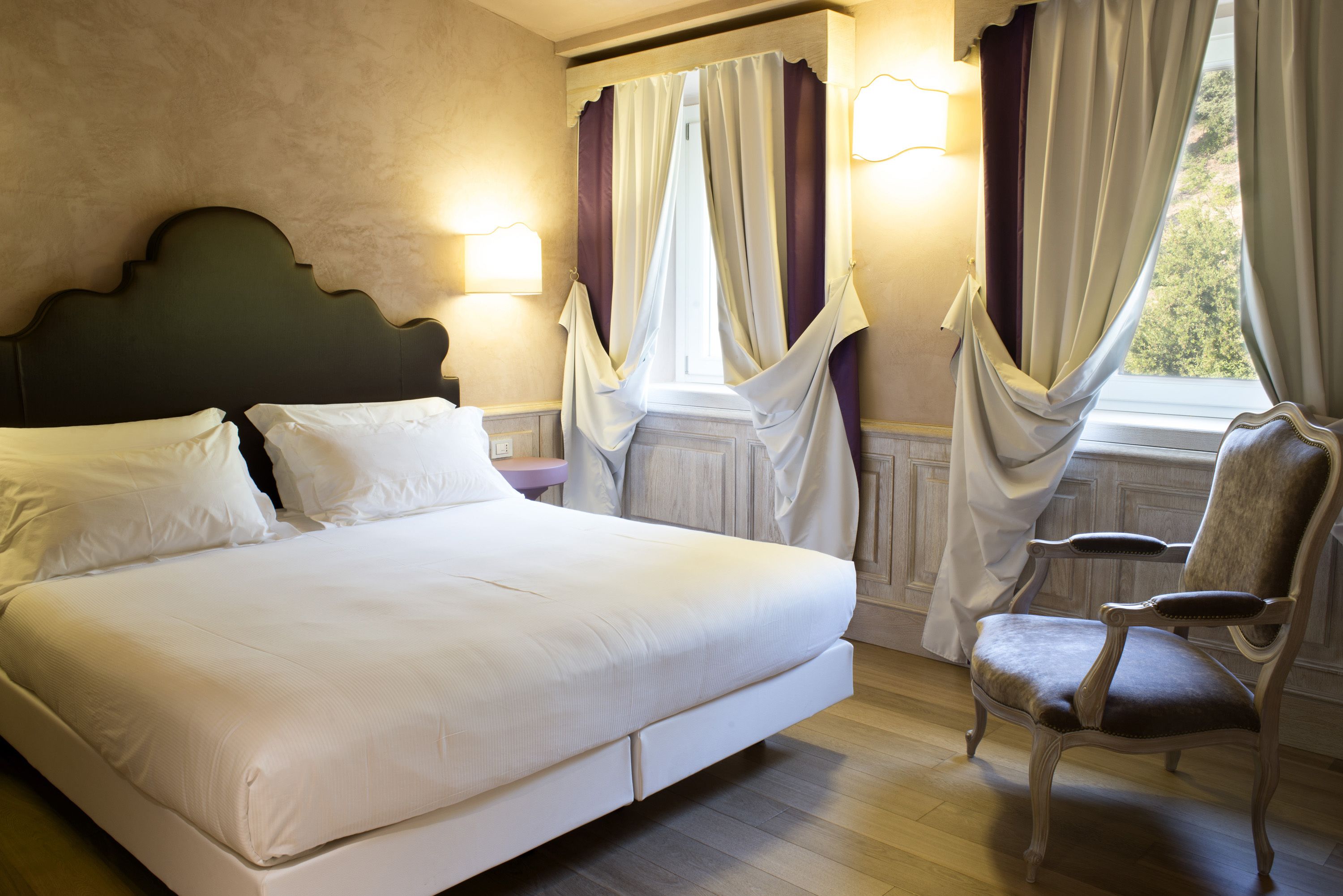 Un lit bien fait, avec du linge de lit blanc, une tête de lit sombre et un plancher en bois. On y trouve une chaise avec un siège et un dossier rembourrés, ainsi qu’une fenêtre dont les rideaux sont partiellement tirés.