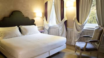 Un lit bien fait, avec du linge de lit blanc, une tête de lit sombre et un plancher en bois. On y trouve une chaise avec un siège et un dossier rembourrés, ainsi qu’une fenêtre dont les rideaux sont partiellement tirés.