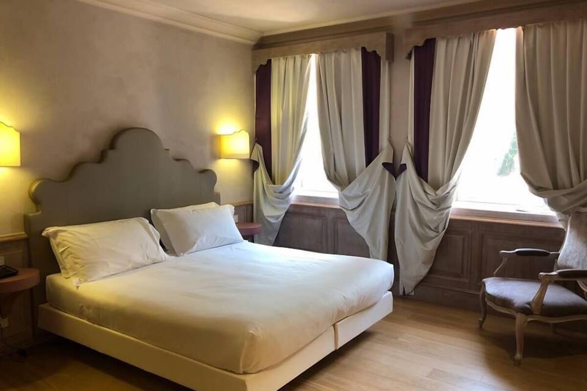 A hotel room with a large bed, two bedside lamps, a chair, and a window with curtains.