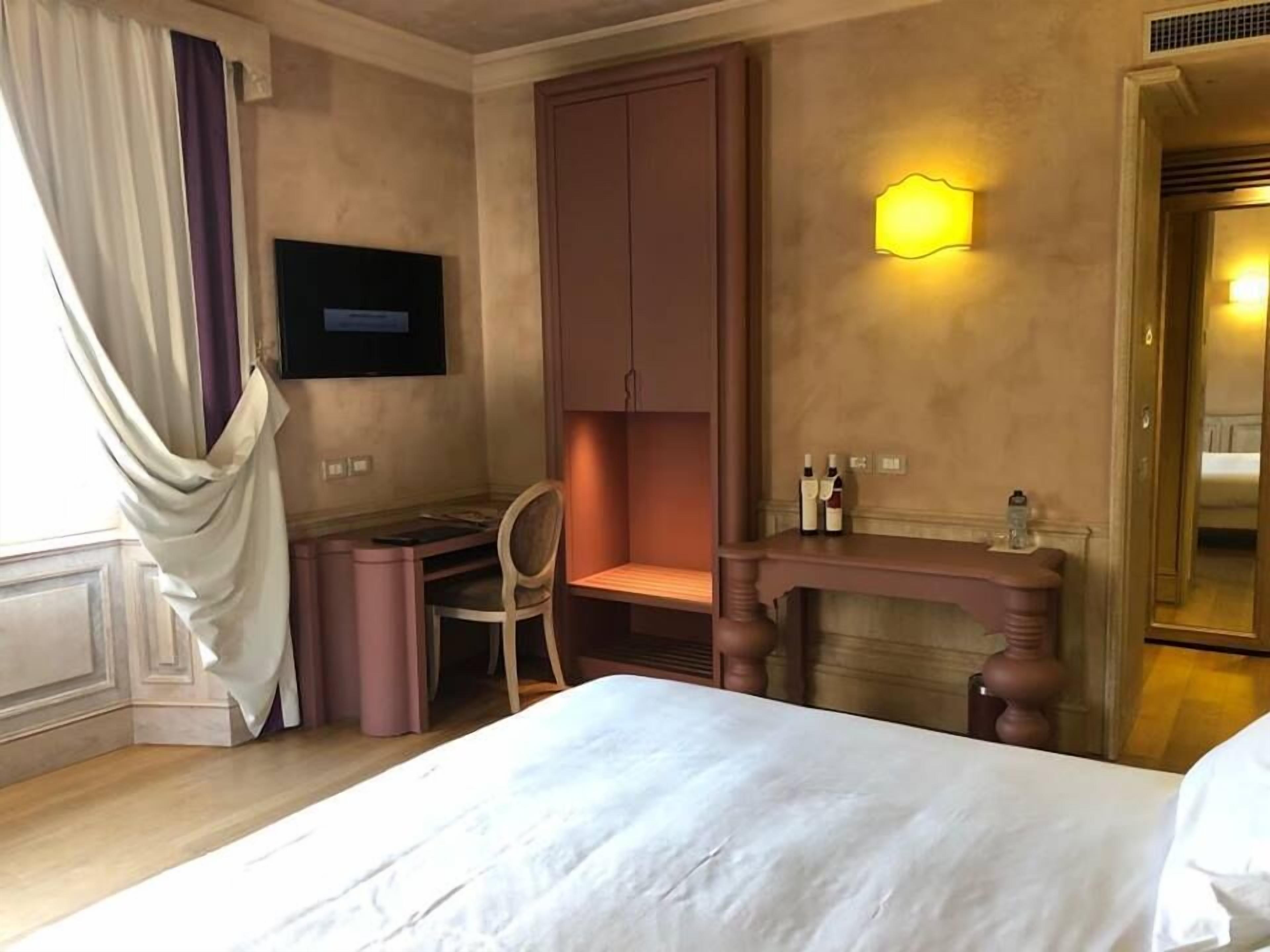 A hotel room with a bed, a desk, a chair, a television, a wall-mounted light, and a fireplace.