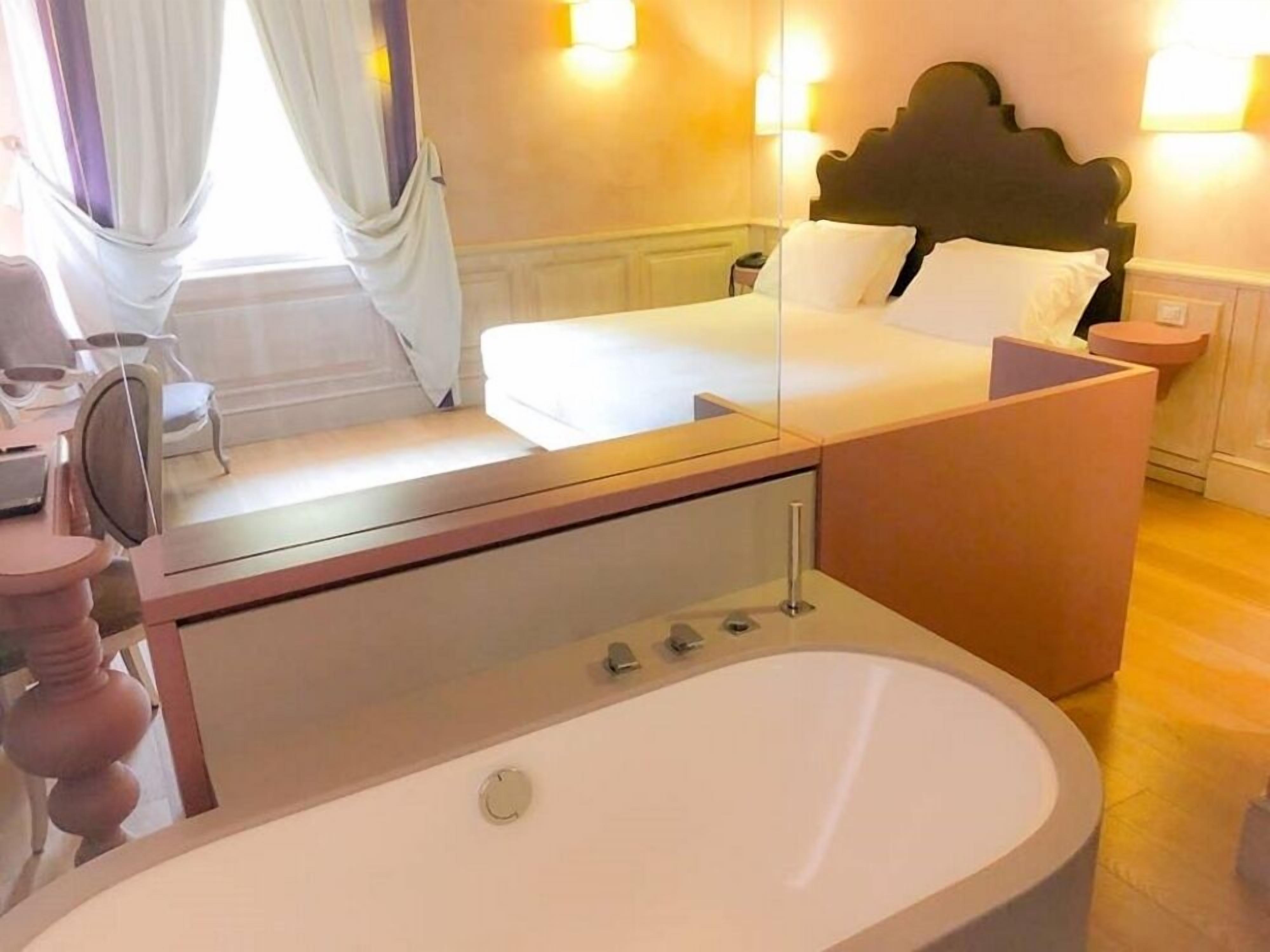 A hotel room with a large bathtub, a bed with white linens, a wooden headboard, and a nightstand with a lamp.