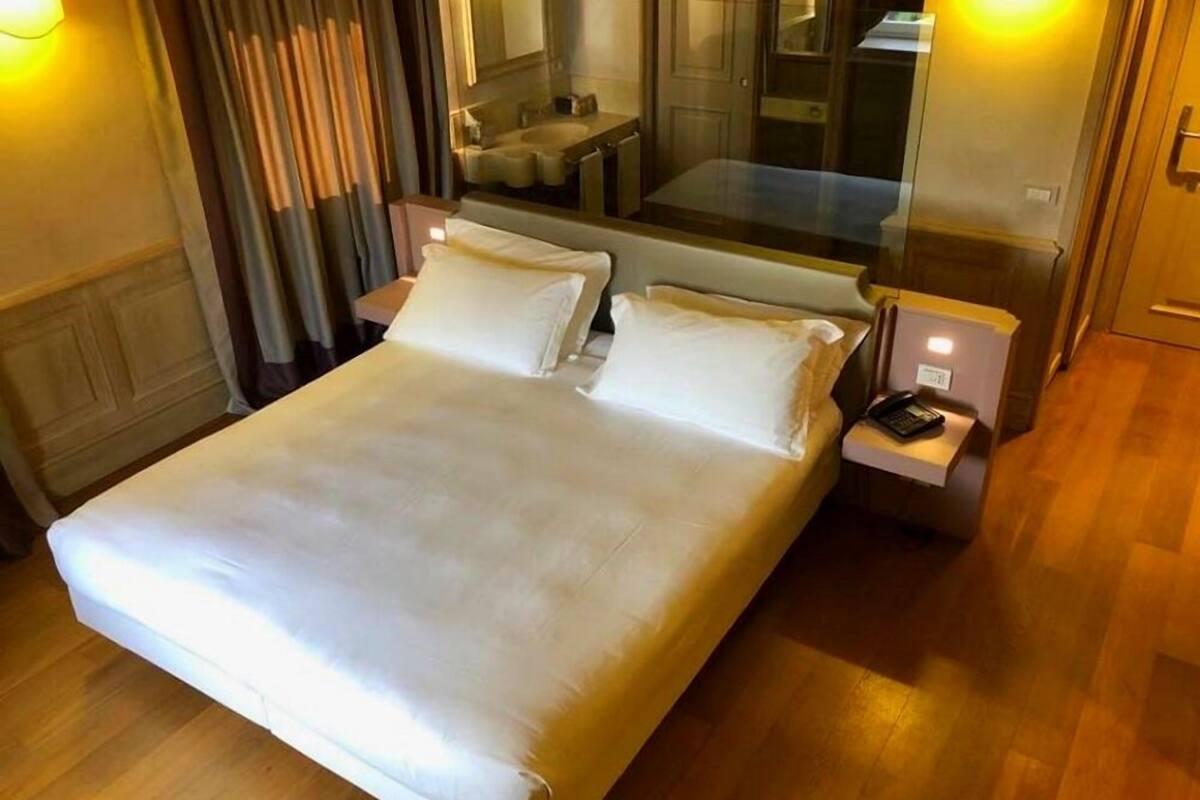 A hotel room with a large bed, bedside tables, a mirror, and a view of a bathroom through a glass partition.