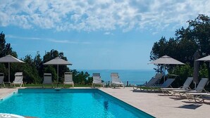 Une piscine entourée de verdure, avec des chaises longues et des parasols, et offrant une vue sur la mer.