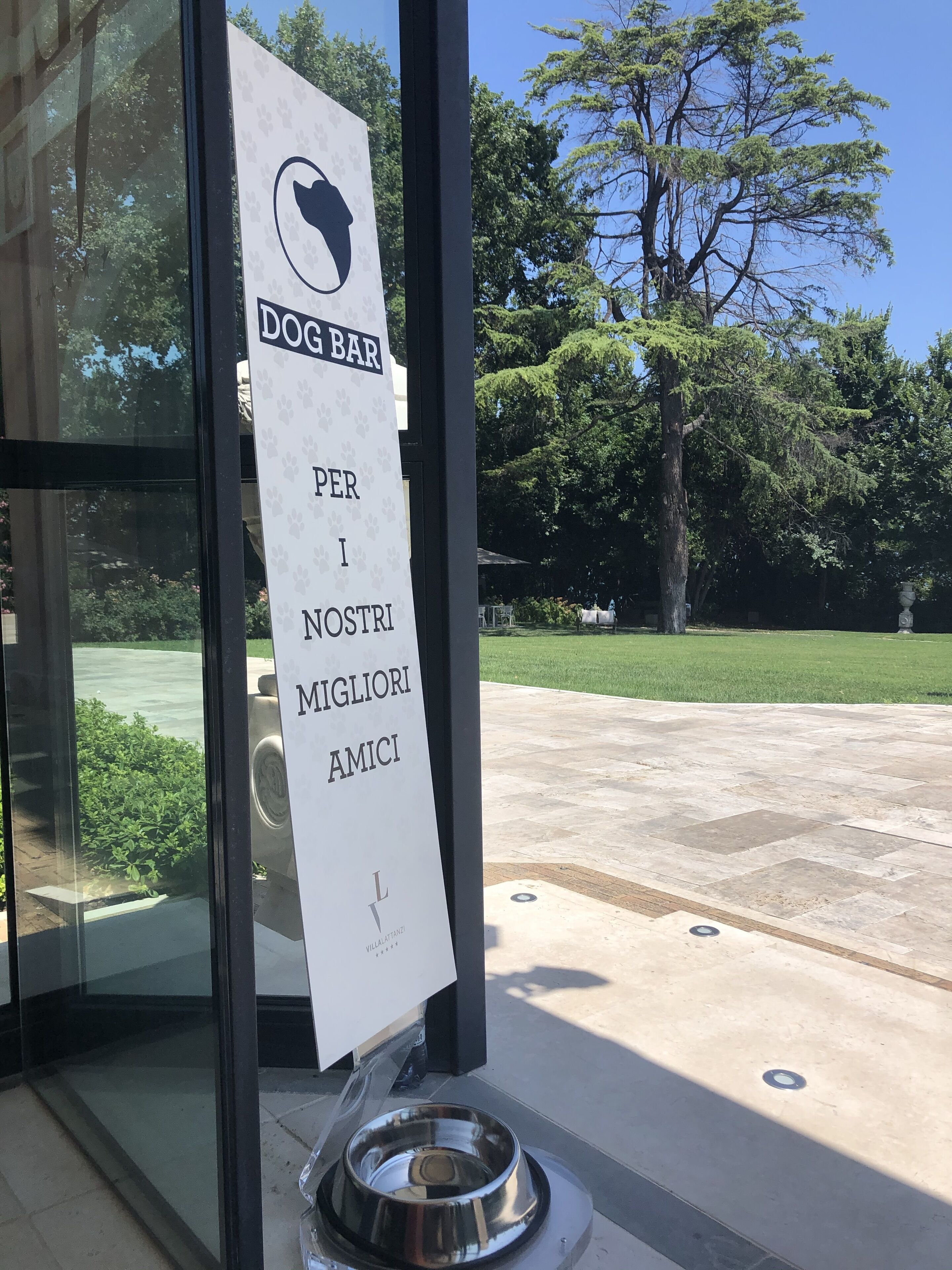 Segnale per un bar per cani all'esterno di un edificio con porta in vetro, una ciotola metallica a terra e un albero sullo sfondo.