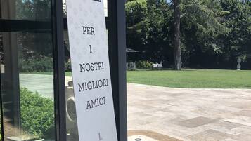 Segnale per un bar per cani all'esterno di un edificio con porta in vetro, una ciotola metallica a terra e un albero sullo sfondo.