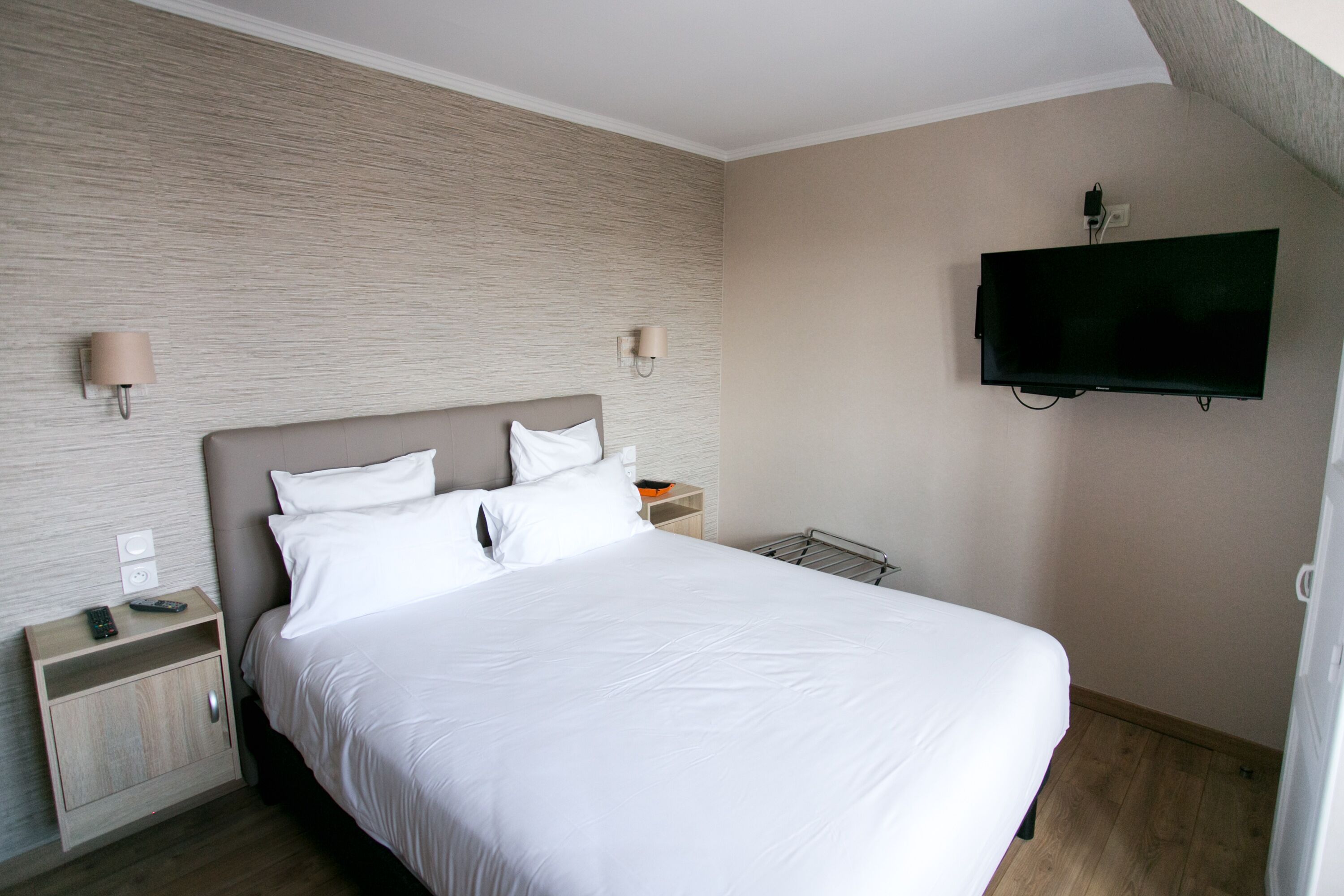 A neatly made bed with white linens, a bedside table with a lamp, and a mounted TV on the wall.