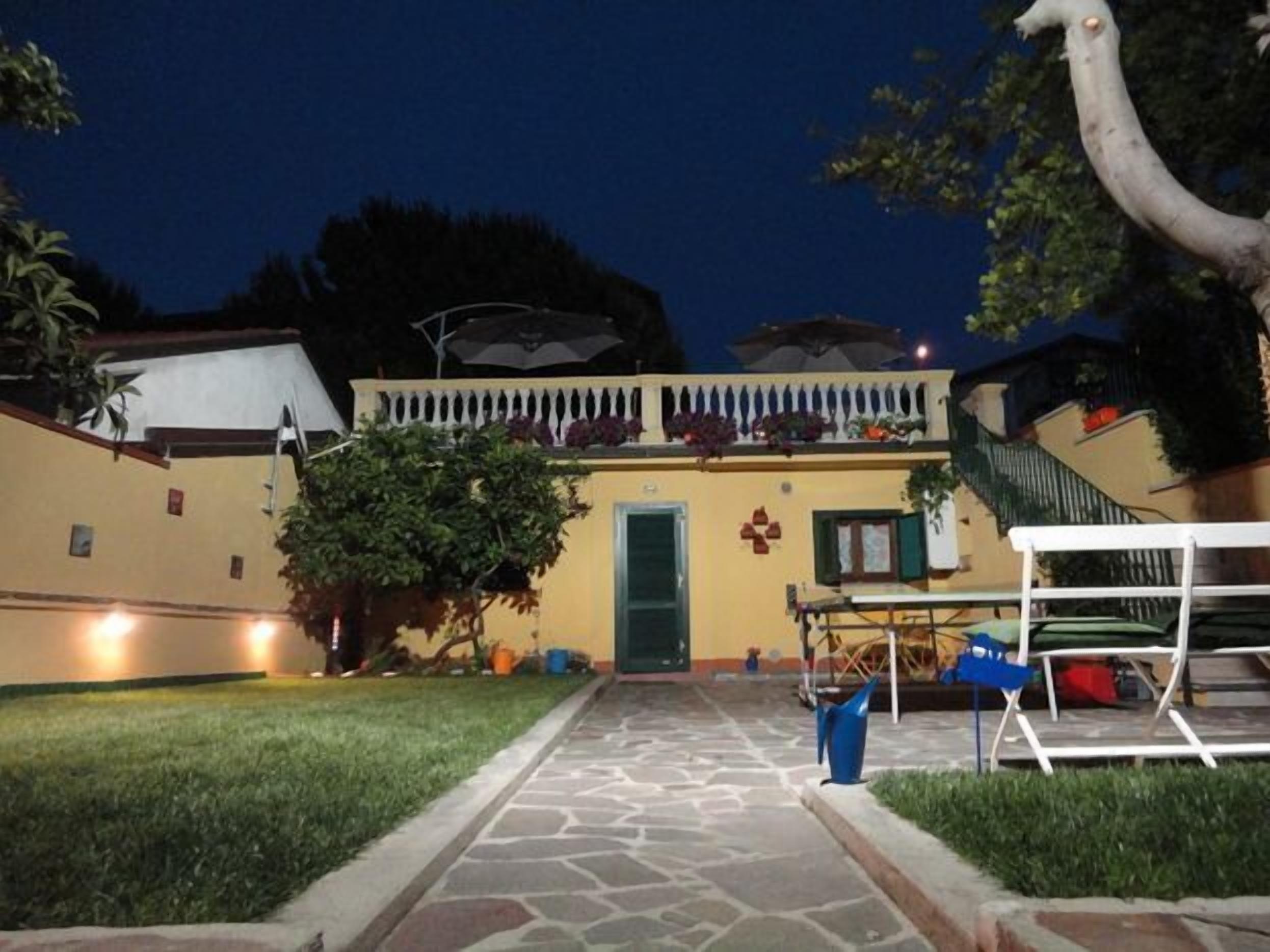 Un cortile con un vialetto in pietra che conduce a un edificio con una porta verde. Si nota una recinzione bianca, un albero e mobili da esterno.