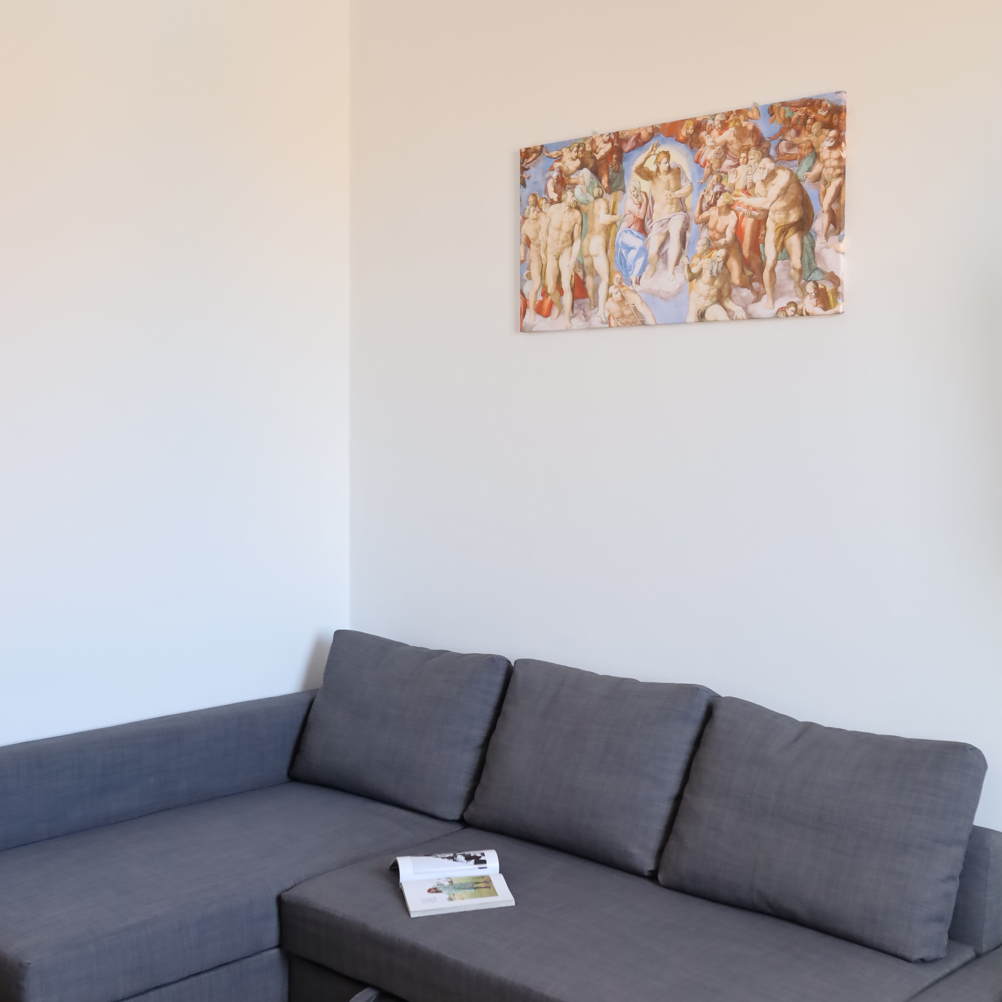 A grey sofa with a magazine on it, and a classical painting on the wall.