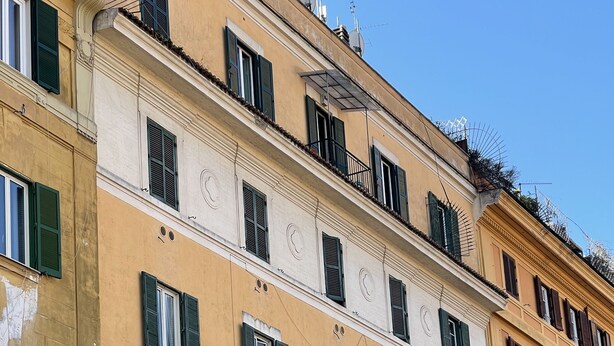 Ein mehrstöckiges Gebäude im klassischen Stil, mit gleichmäßig angeordneten Fenstern mit grünen Fensterläden, einem Balkon im zweiten Stock und blauem, wolkenlosem Himmel im Hintergrund.