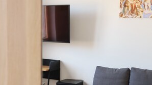 Ein Zimmer mit grauem Sofa, einem schwarzen Fernseher an der Wand, einem kleinen schwarzen Tisch und einem Gemälde.