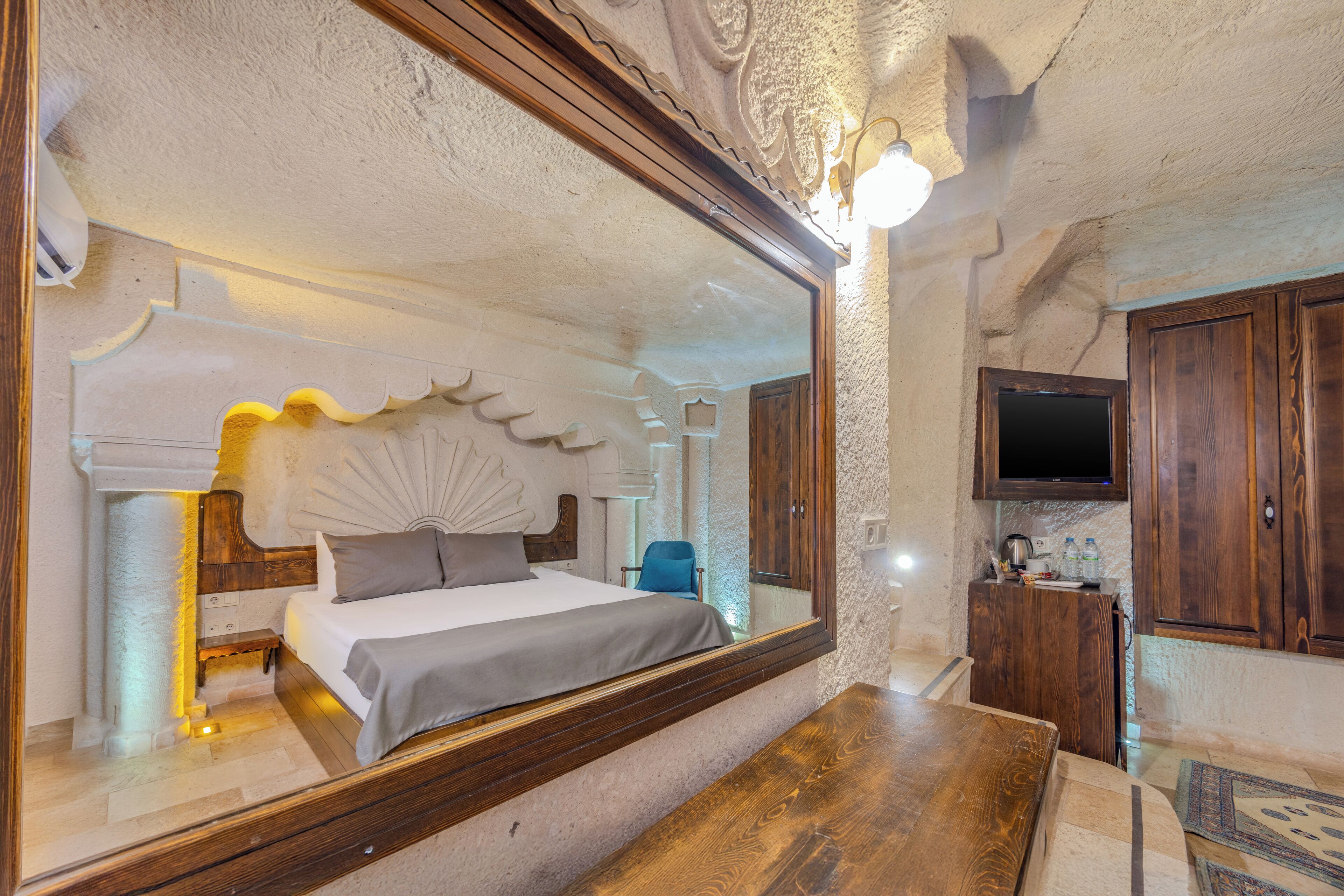 A bedroom with a large mirror, a bed with a headboard, a wooden bench, a TV, and a small kitchenette.
