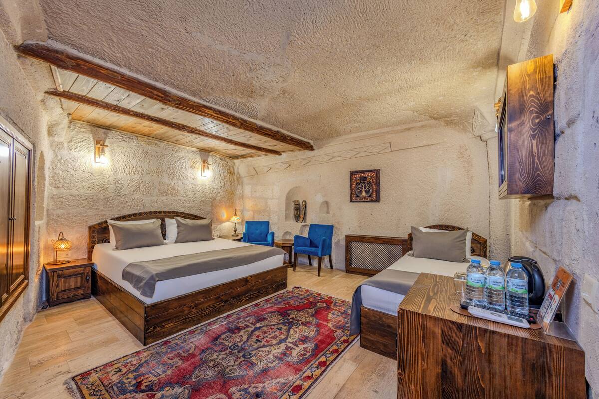 A cozy room with a large bed, two smaller beds, a wooden table with water bottles, a red patterned rug, and a stone wall.