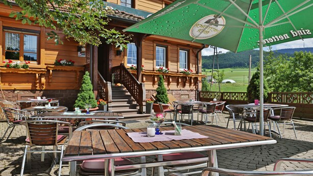 An outdoor patio with wooden tables and chairs, a green umbrella, and a view of a green field in the background. - Grüner Hof (Zell am Harmersbach)