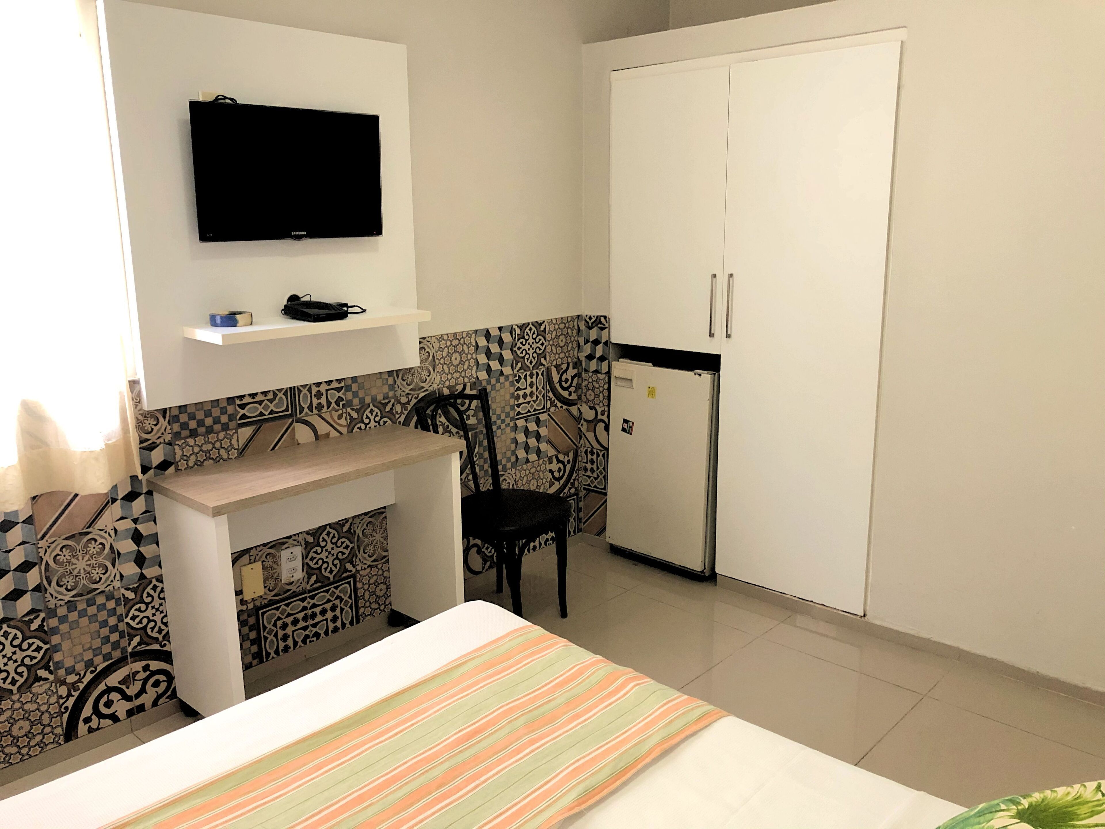 A small, clean room with a bed, a chair, a TV mounted on the wall, a white wardrobe, and a refrigerator.