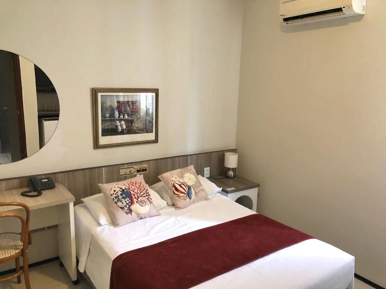 A hotel room with a bed, a desk, a chair, a nightstand, a lamp, and an air conditioning unit.