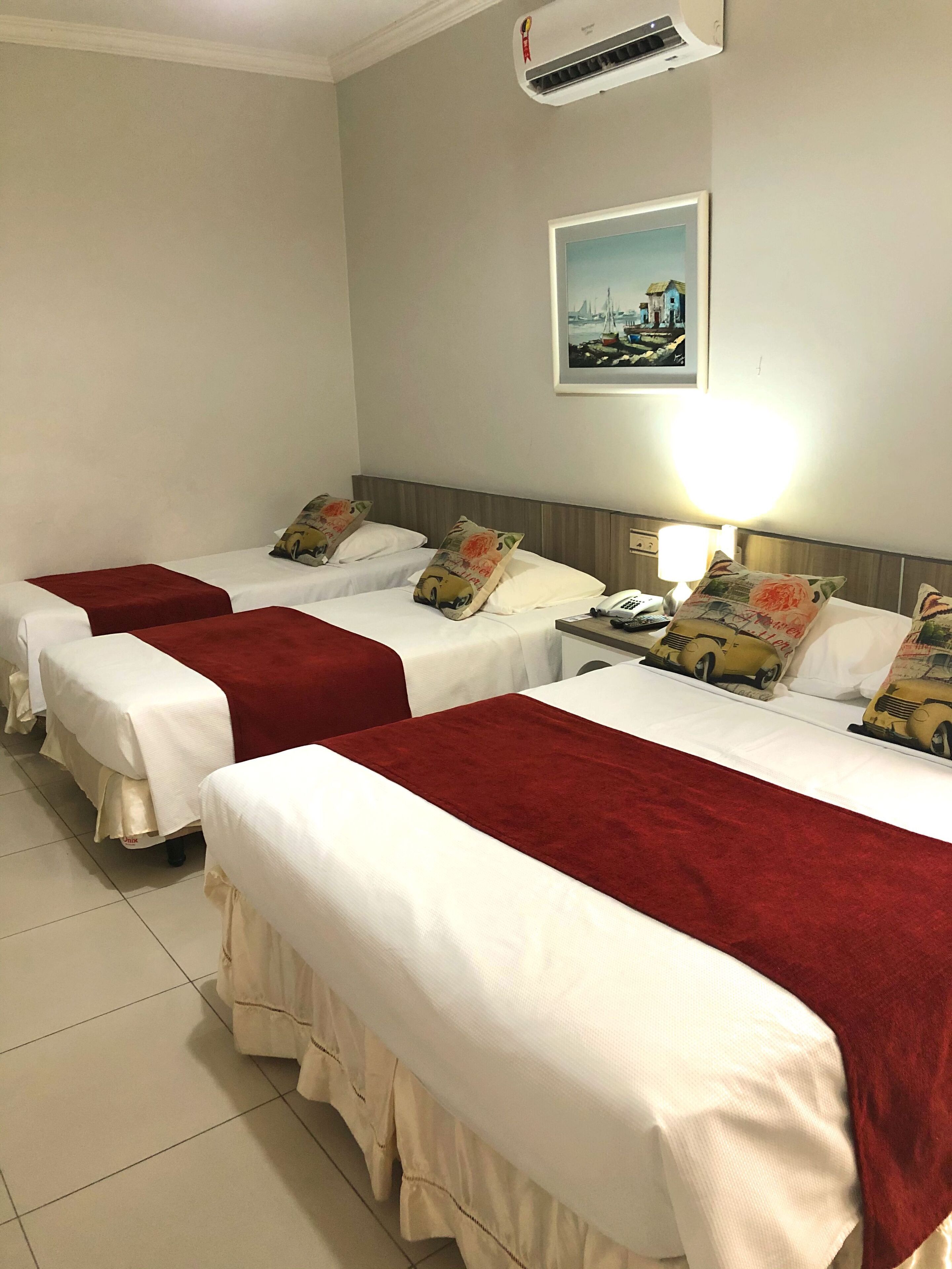 A hotel room with three single beds, each with a red blanket and white bedding. There is a wall-mounted air conditioner and a framed picture above the beds.