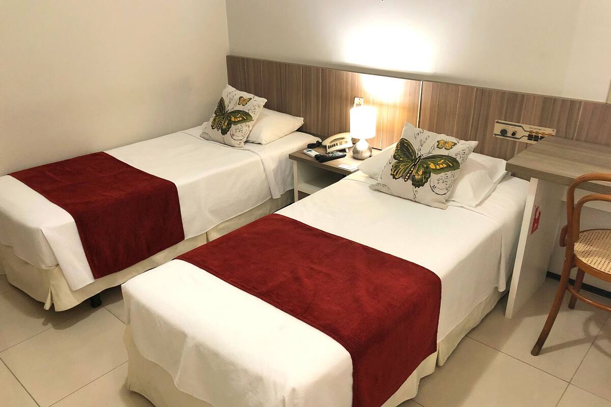 A hotel room with two single beds, each with a red blanket and a pillow with a butterfly design. There is a wooden nightstand with a lamp, a small table with a telephone, and a wooden chair.