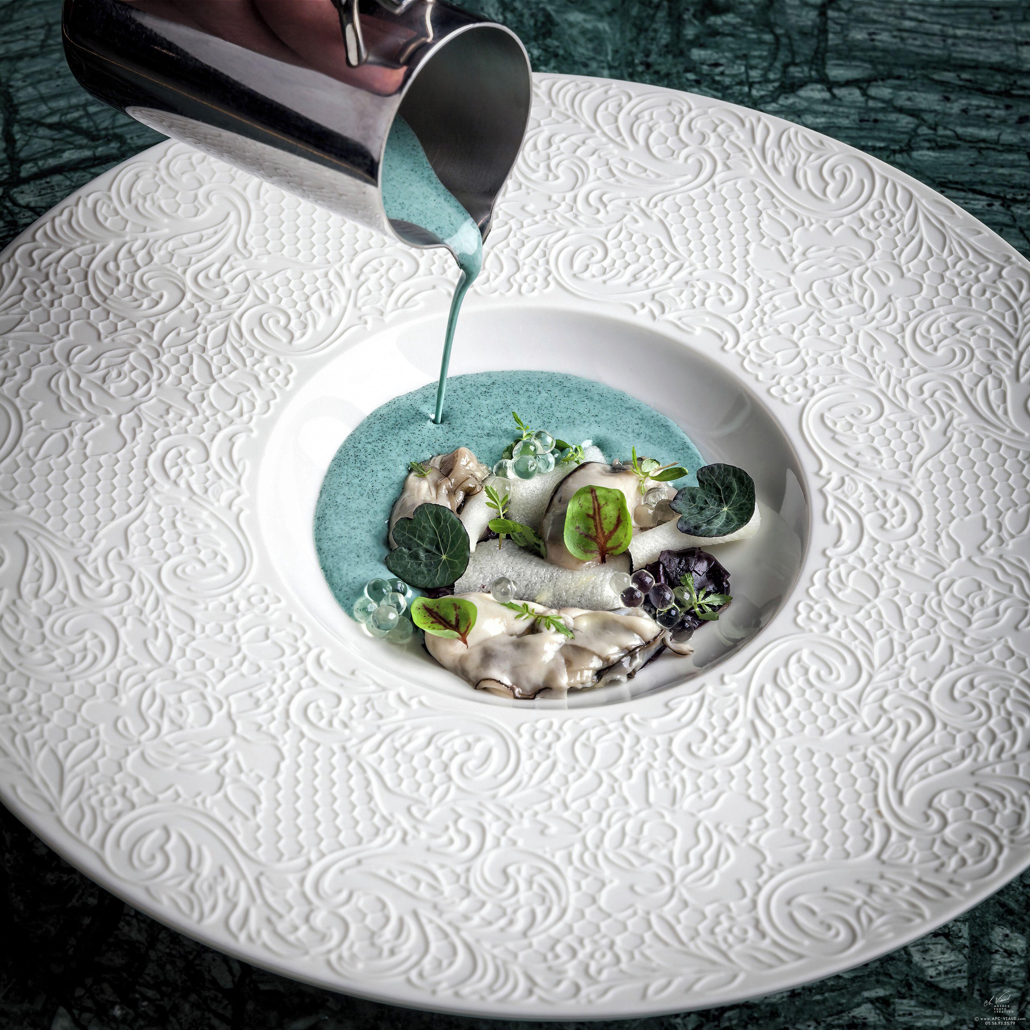 Un plato blanco con diseños estampados elaborados, salsa azul siendo servida y diversos adornos, incluyendo vegetales verdes y champiñones.