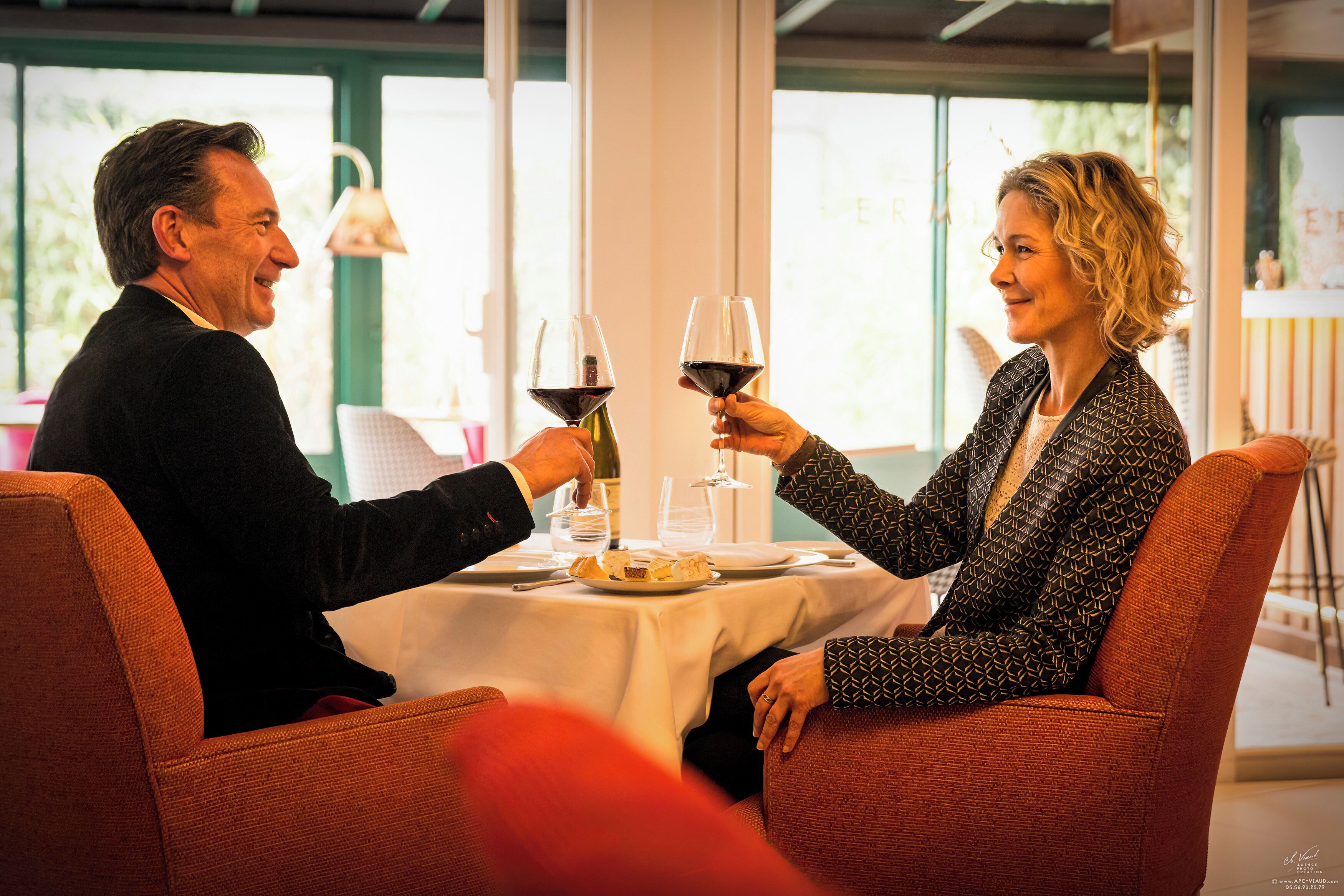 Un couple est assis à une table, devant des verres à vin, une assiette de nourriture, et une fenêtre donnant sur le paysage.