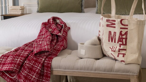Een net opgemaakt bed met een witte deken, groene kussens, een rood-witte gebloemde plaid en een beige plaid, plus een beige tas met rode tekst.