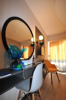 Habitación de hotel con un espejo grande, un escritorio con una silla, una escultura de vidrio decorativa y vista del cuarto a través de una ventana con cortinas.