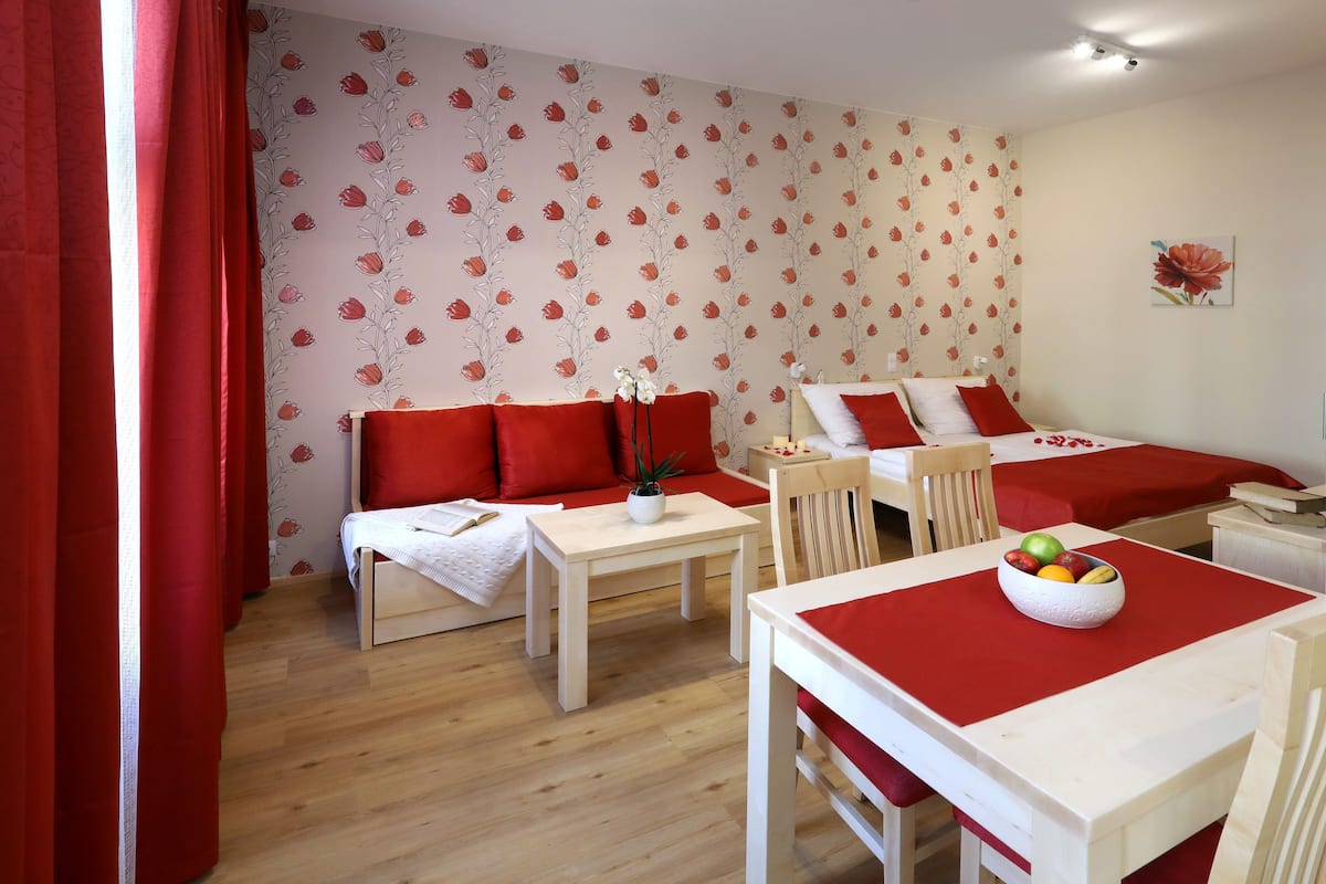 A room with a bed, a small table with a bowl of fruit, a chair, and a sofa with red cushions.