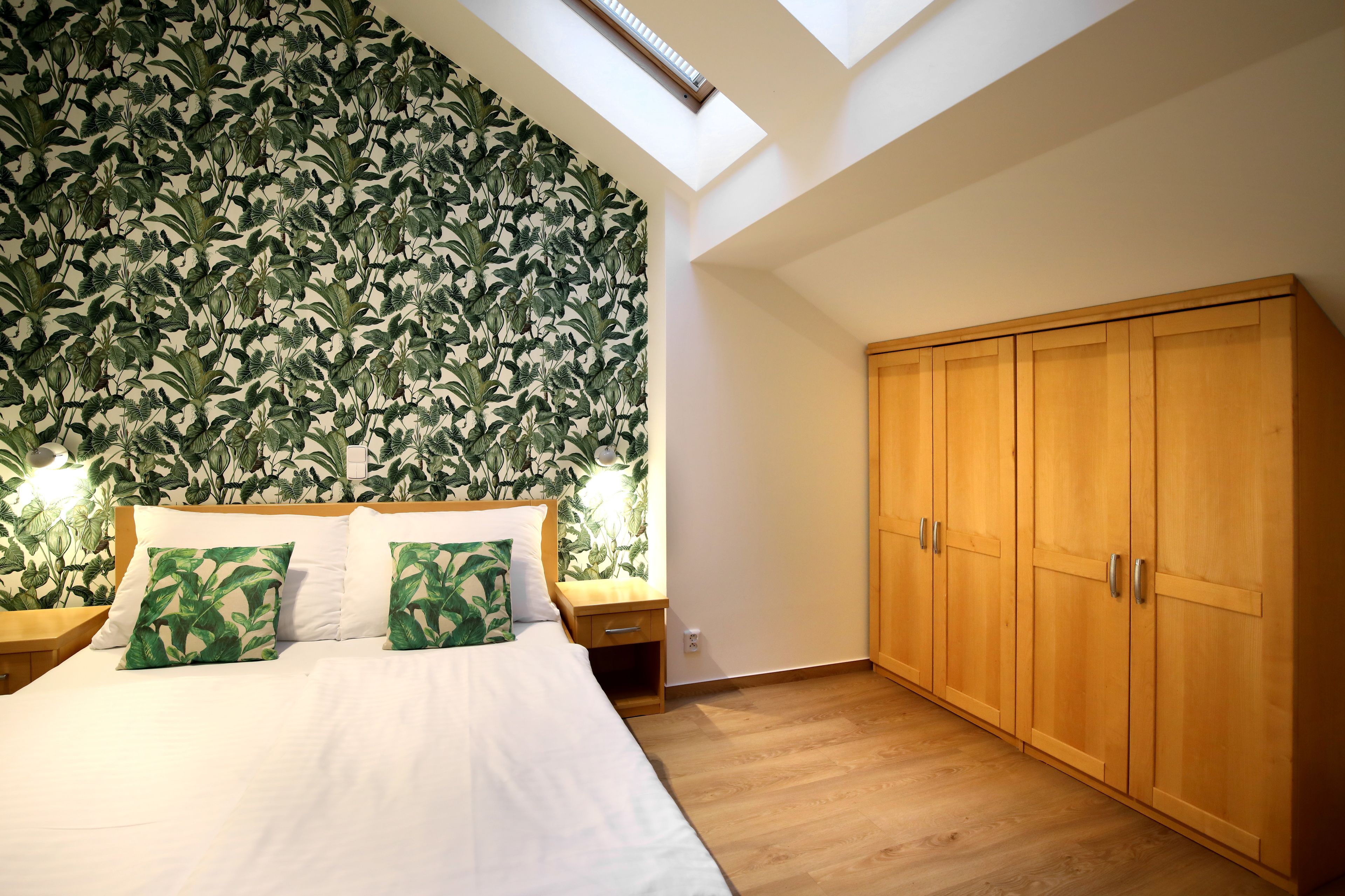 Une chambre avec un puits de lumière, un lit avec une literie blanche et des oreillers à motifs de feuilles vertes, une armoire en bois et une table de chevet avec une lampe.