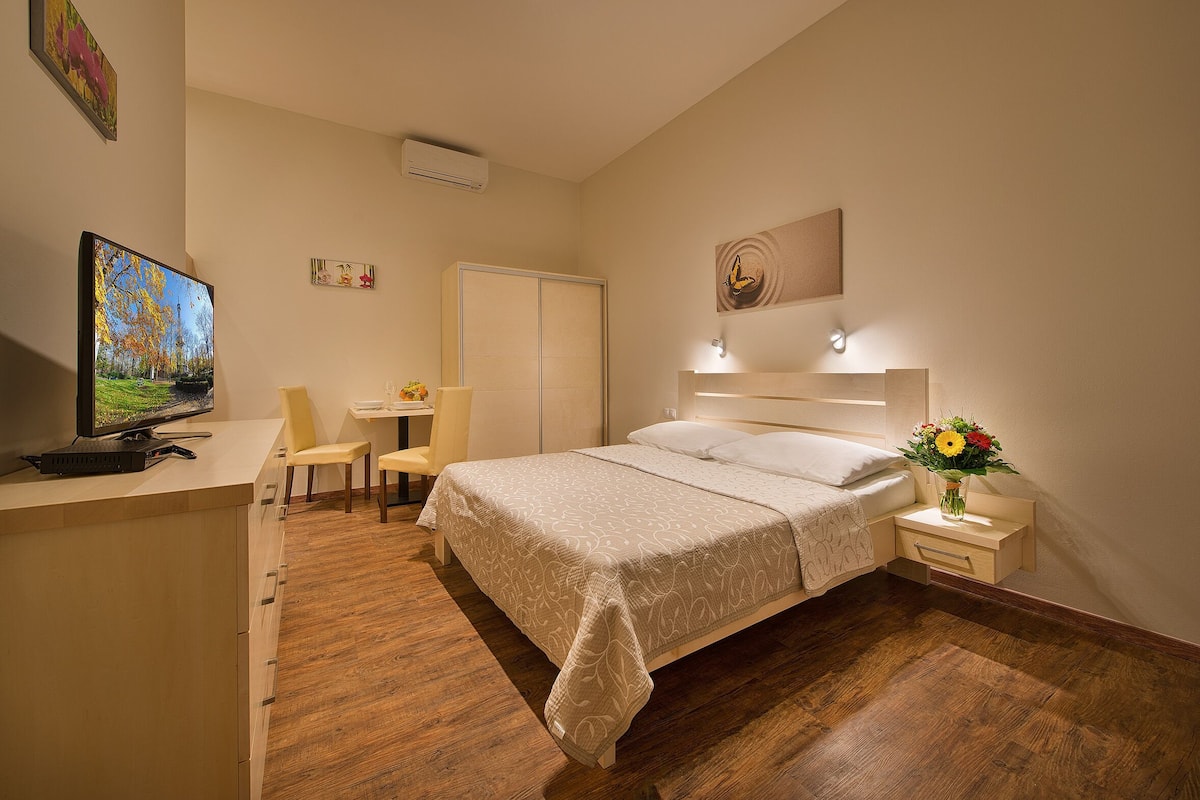A bedroom with a bed, bedside table, TV, desk, chair, and a small wall-mounted shelf.