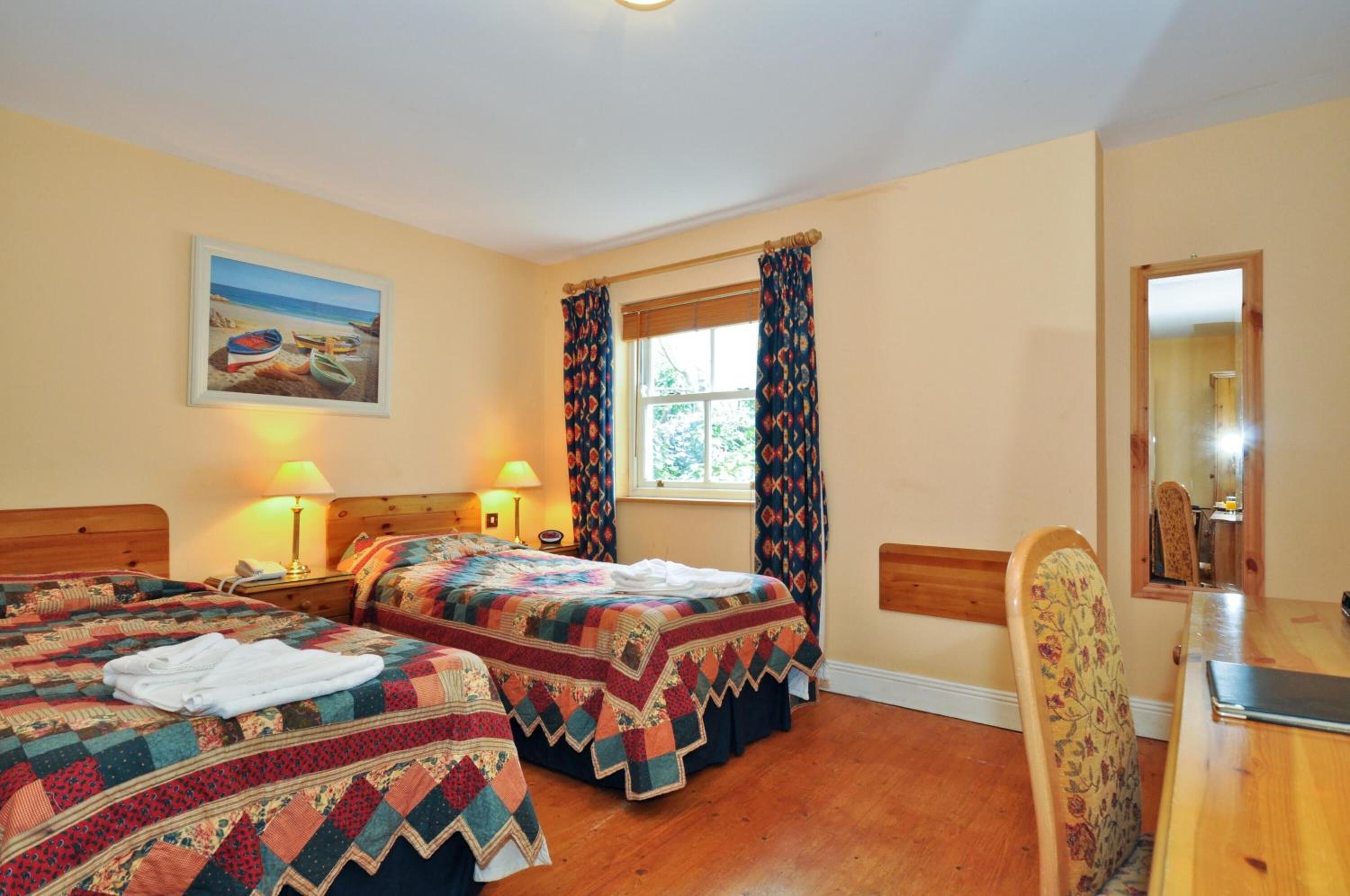 Une chambre d’hôtel avec deux lits, une tête de lit en bois, une fenêtre avec des rideaux, un tableau au mur et une table à manger en bois avec une chaise.
