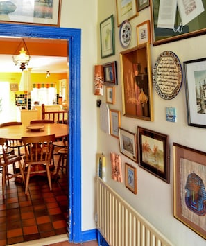 Um corredor com uma porta azul que dá para uma sala de jantar com cadeiras de madeira e uma mesa. As paredes são decoradas com diversas molduras e obras de arte.