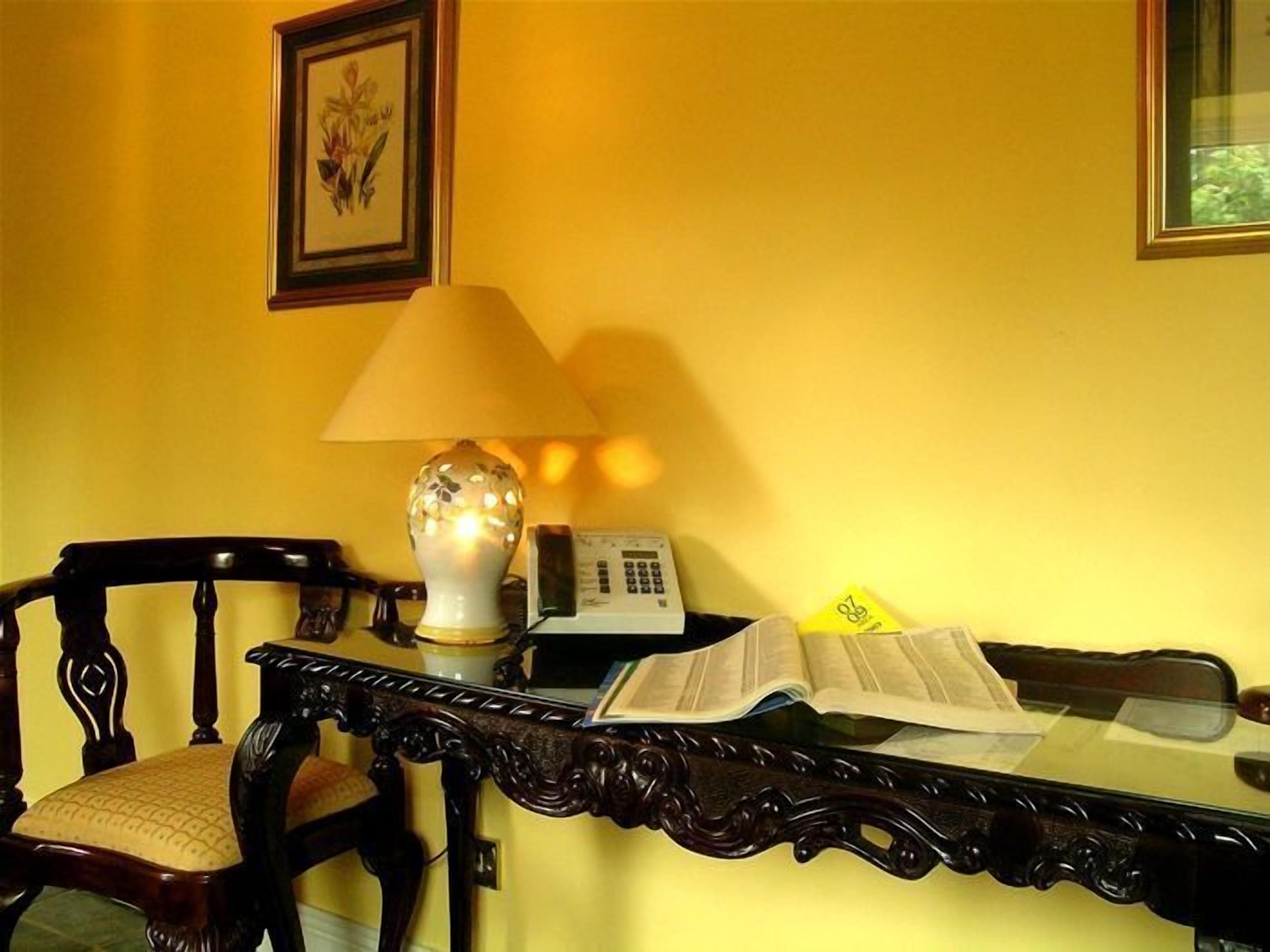 A room with a yellow wall, a wooden chair, a table with a lamp, a telephone, and an open book.
