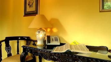 A room with a yellow wall, a wooden chair, a table with a lamp, a telephone, and an open book.