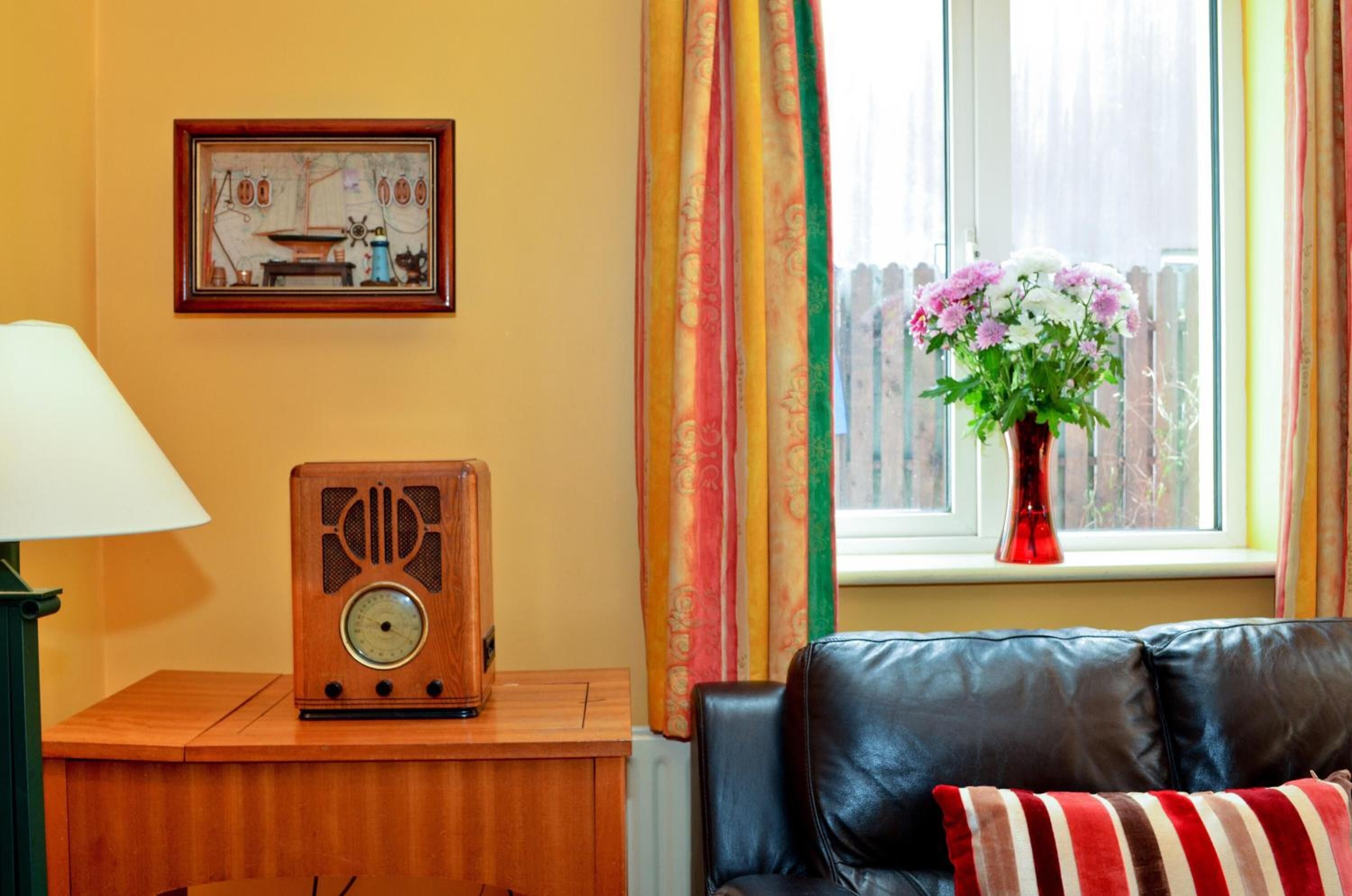A room with a wooden radio on a table, a black leather sofa, a window with curtains, and a vase with flowers on the windowsill.