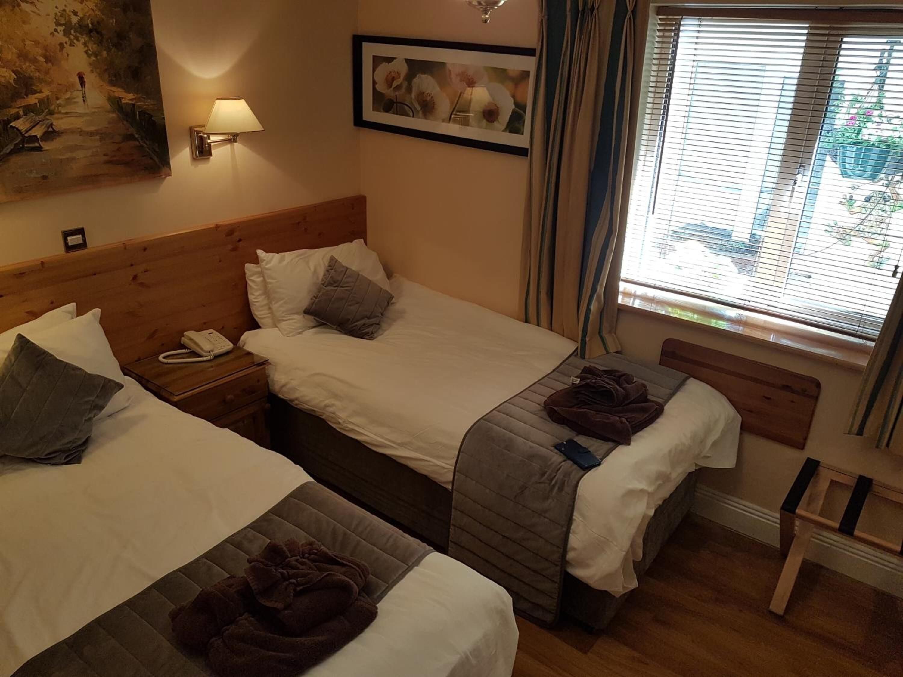 A hotel room with two beds, a bedside table with a phone, a lamp, and a window with curtains.