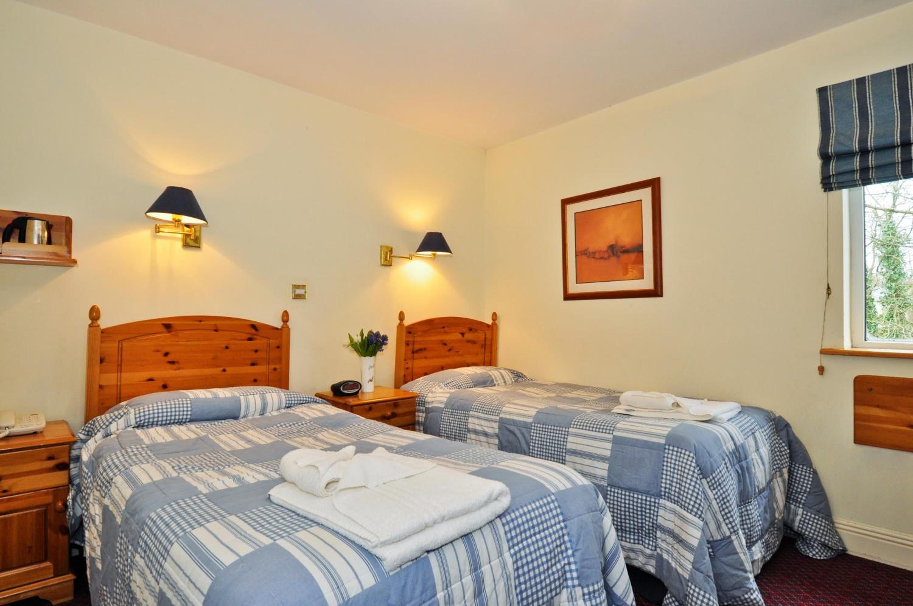 A bedroom with two beds, wooden headboards, bedside tables, lamps, a framed picture, and a window with a view.