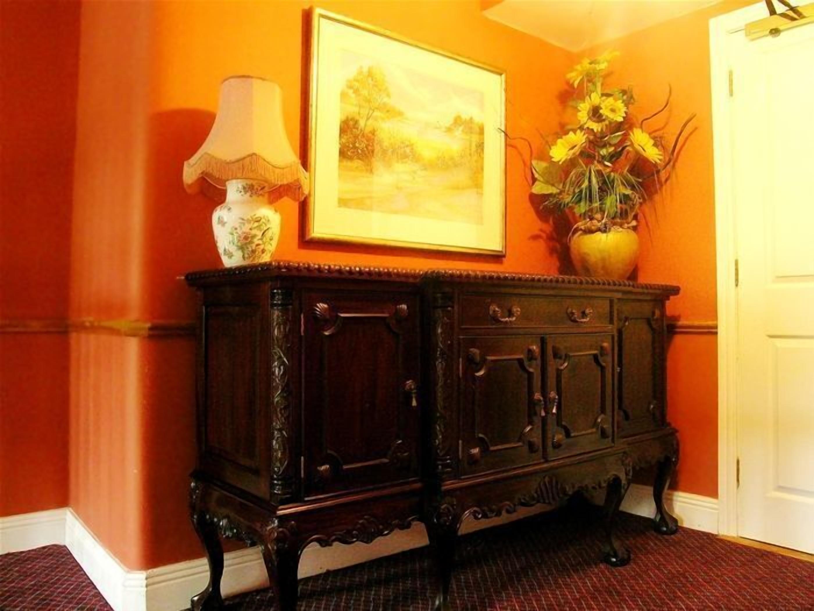 Un mueble de madera oscura con intrincados adornos, un cuadro de paisaje enmarco y un jarrón con girasoles sobre un suelo cubierto de moqueta roja.