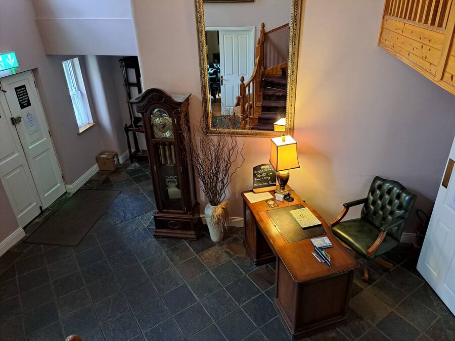 A room with a wooden desk, a green leather chair, a tall grandfather clock, a mirror, and a staircase.