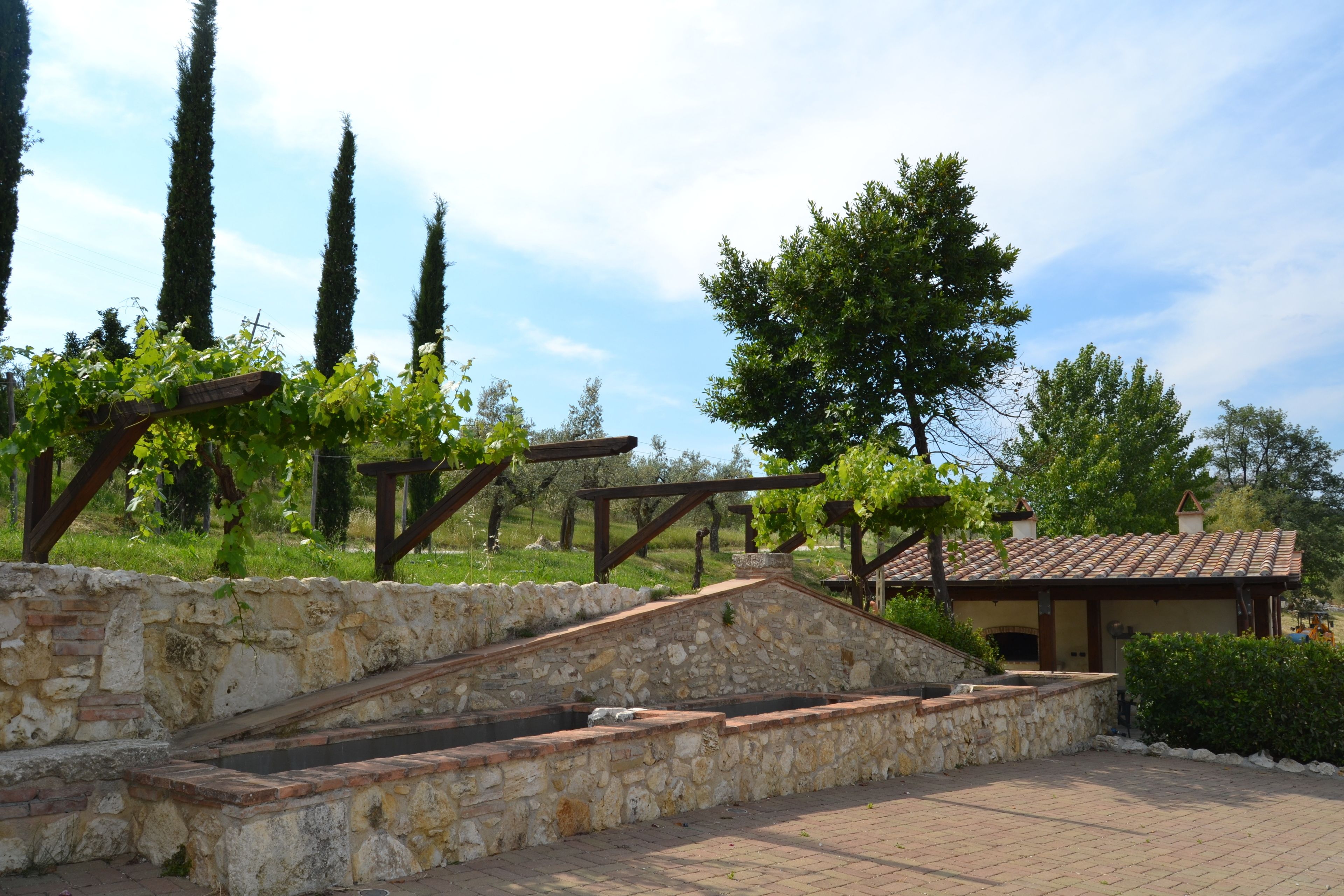 Un muro di sostegno in pietra con una scala in pietra, una pergola con viti e un edificio con tetto in tegole.