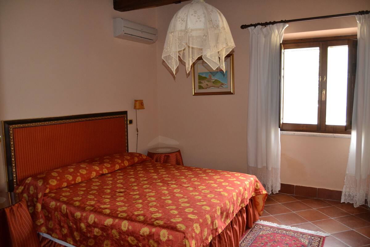 A bedroom with a double bed, a small bedside table, a lamp, a window with curtains, and a painting on the wall.