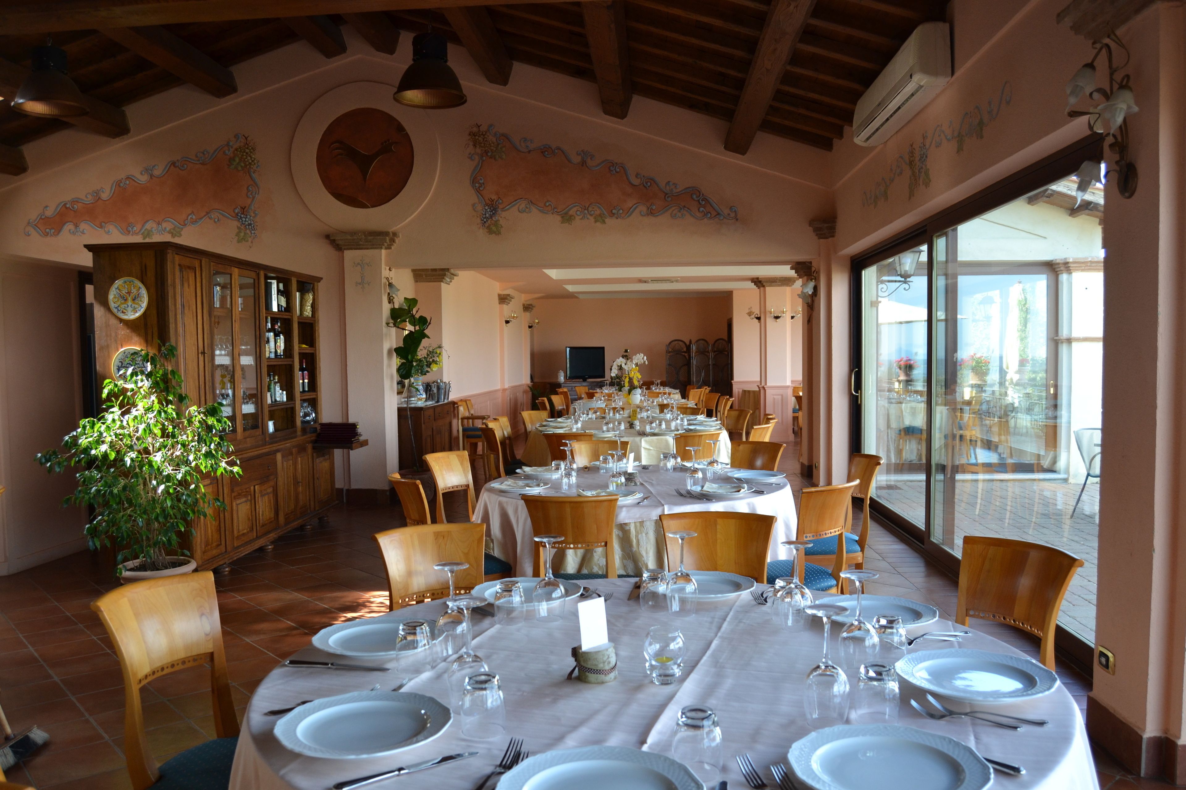 A dining area with round tables set for a meal, wooden chairs, a potted plant, and a view through large windows.