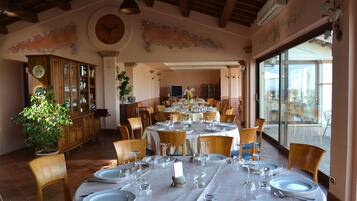 A dining area with round tables set for a meal, wooden chairs, a potted plant, and a view through large windows.