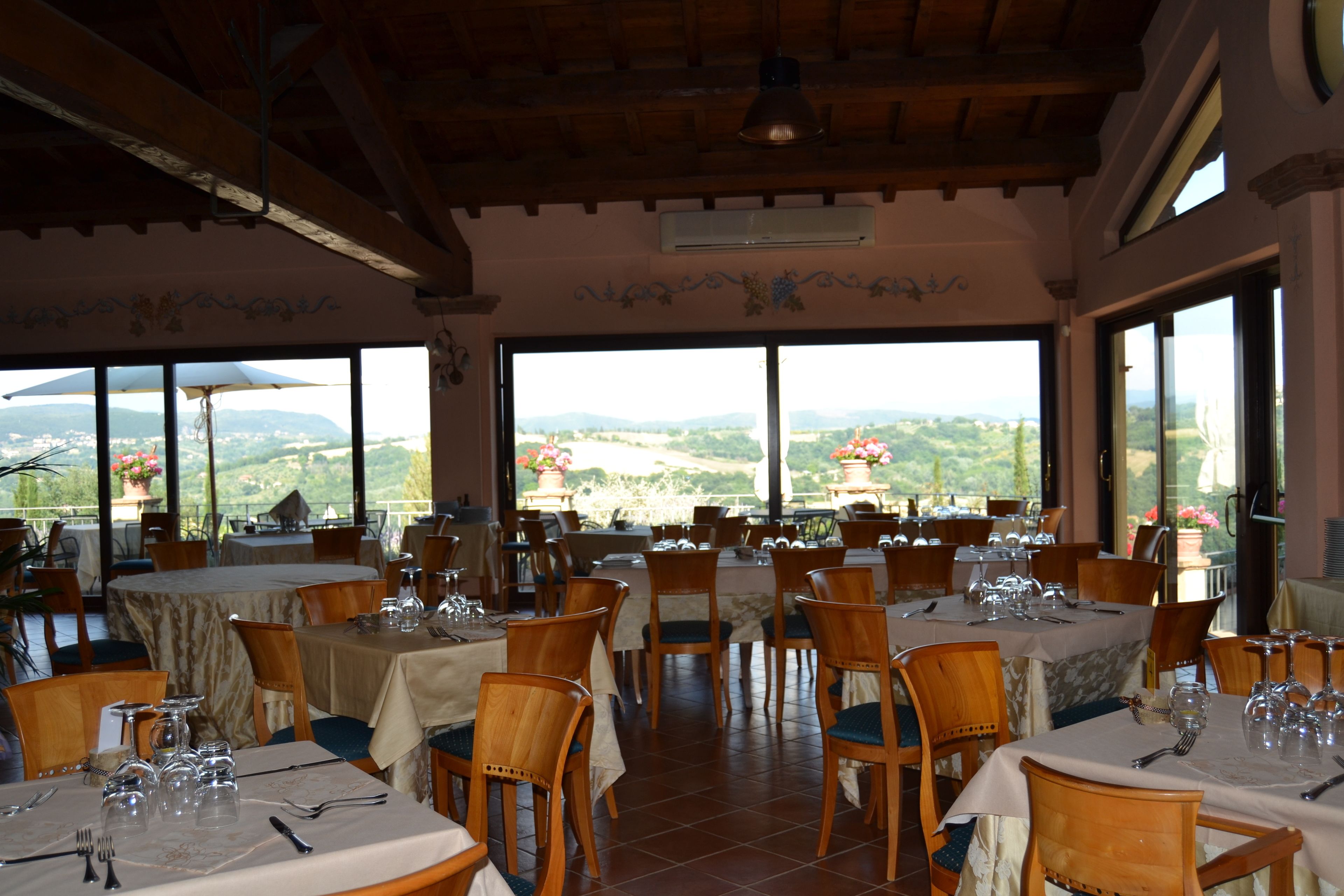 A restaurant with multiple tables set for dining, large windows offering a view of the countryside, and a wooden ceiling.