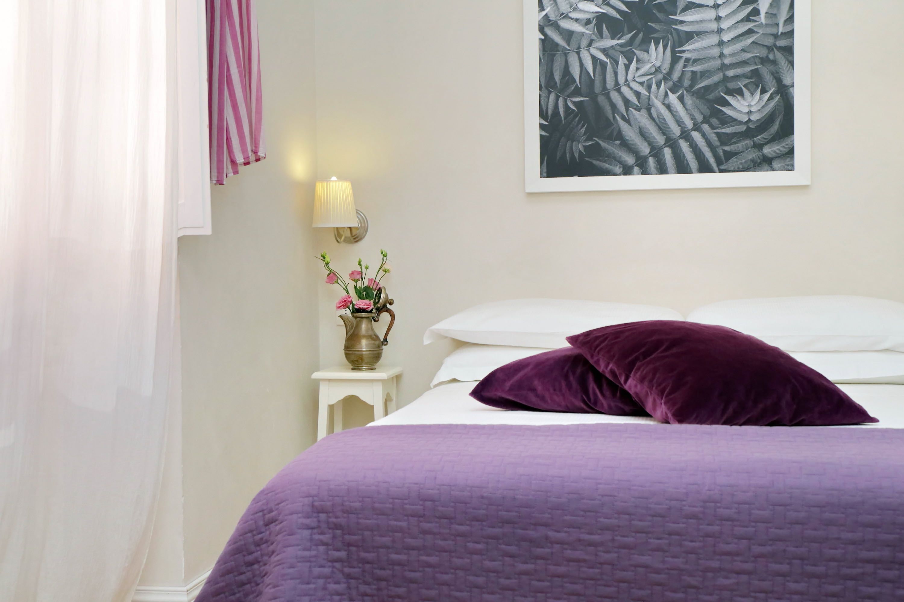 Ein ordentlich bezogenes Bett mit lila Bettdecke, eine Vase mit rosa Blumen, eine Wandlampe und ein gerahmtes Pflanzenbild an der Wand.