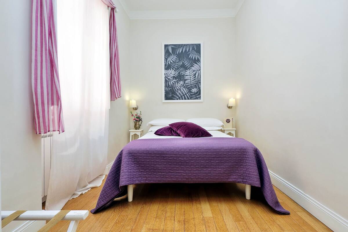 A bedroom with a bed, purple bedding, a wall-mounted artwork, bedside lamps, and a window with curtains.