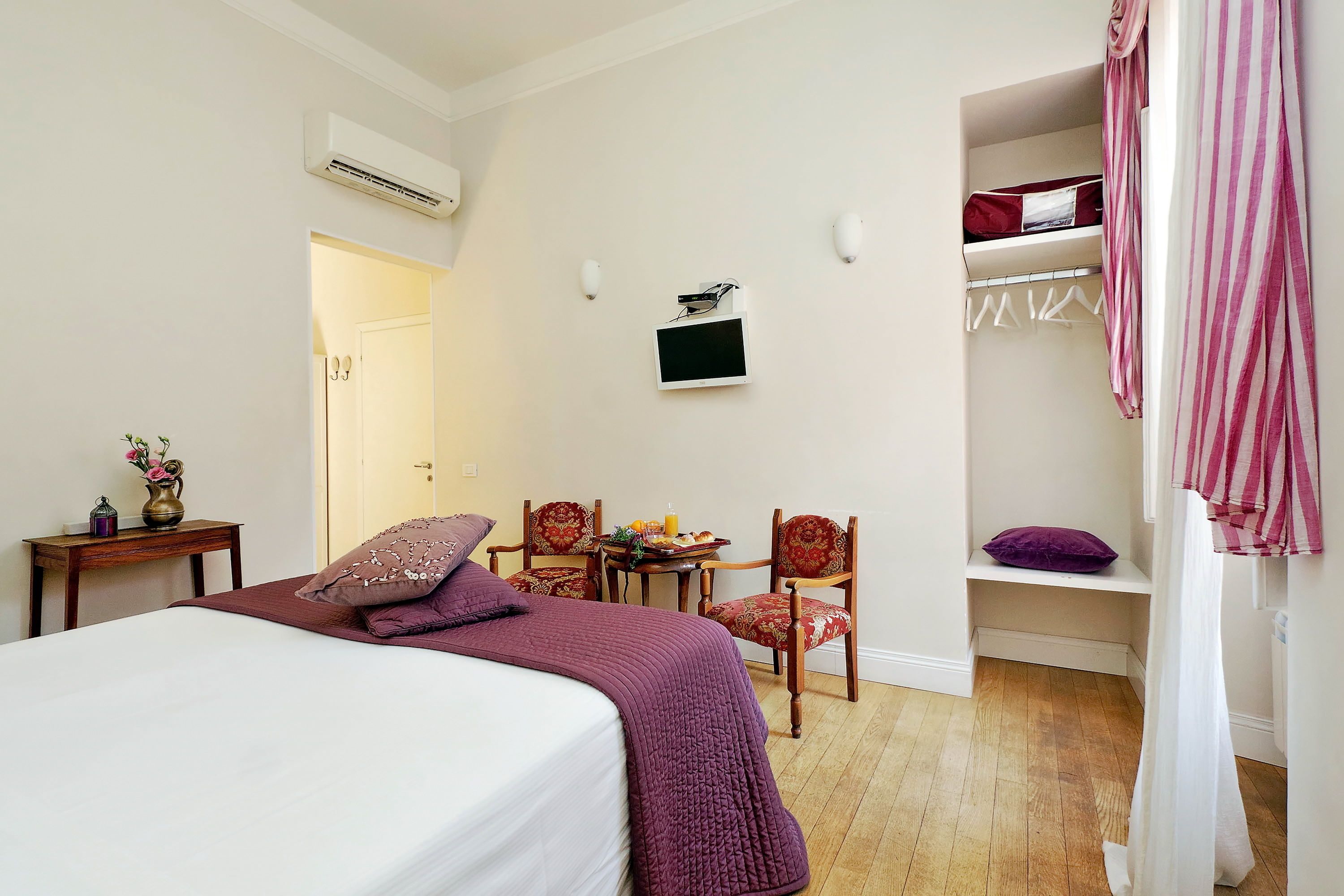 A bedroom with a bed, bedside table, chairs, a small table with items, a TV, and a closet with hanging clothes.
