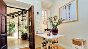 A room with a wooden floor, a white desk with a vase of flowers, a clock on the wall, and a door leading to a balcony with potted plants. - Amaranto Romano (Rome)