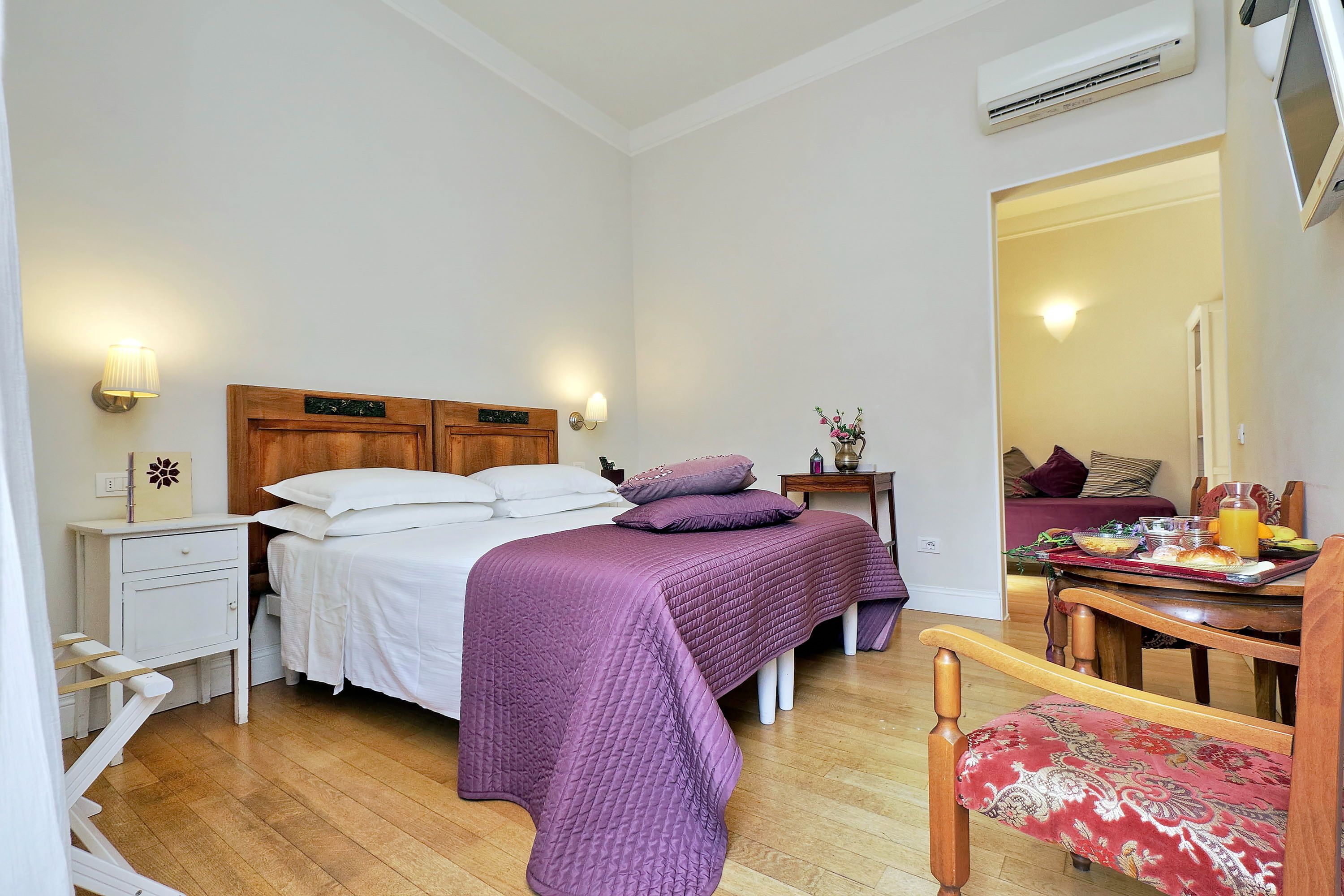 A bedroom with a bed, bedside tables, a chair, a dining table with breakfast items, and a hallway leading to another room.